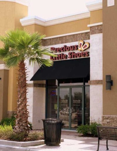 Storefront of "precious little shoes" with a palm tree and bench outside.