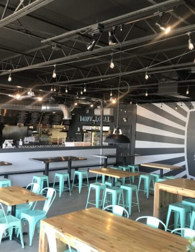 Interior of a modern cafe with empty tables and chairs, featuring an open kitchen and industrial-style ceiling.