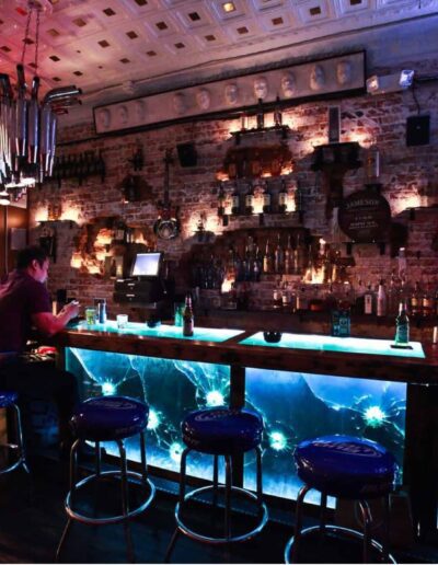 Dimly lit bar with patrons seated, illuminated counter, and televisions displaying content.