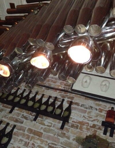 Exposed metal pipes repurposed as light fixtures above a row of wine bottles mounted on a brick wall.