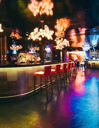 An empty nightclub with colorful lighting, bar stools lined up, and ornate chandeliers.