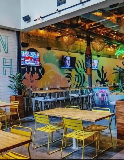 Interior of a vibrant restaurant with neon signage, tropical wallpaper, and yellow chairs.